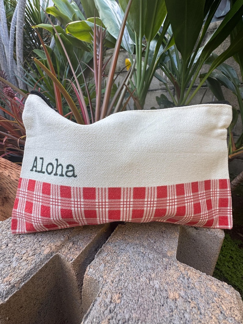 May include: Cream-colored rectangular pouch with a red and white plaid pattern at the bottom. The word "Aloha" is printed in black on the upper portion of the pouch. The pouch has a black zipper.