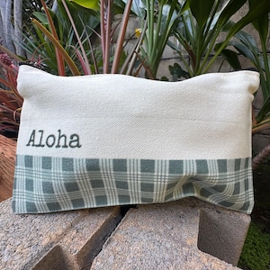 May include: Cream-colored rectangular pouch with the word "Aloha" embroidered in black. The bottom section features a green and white plaid pattern. The pouch is resting on a textured surface, with lush greenery in the background.