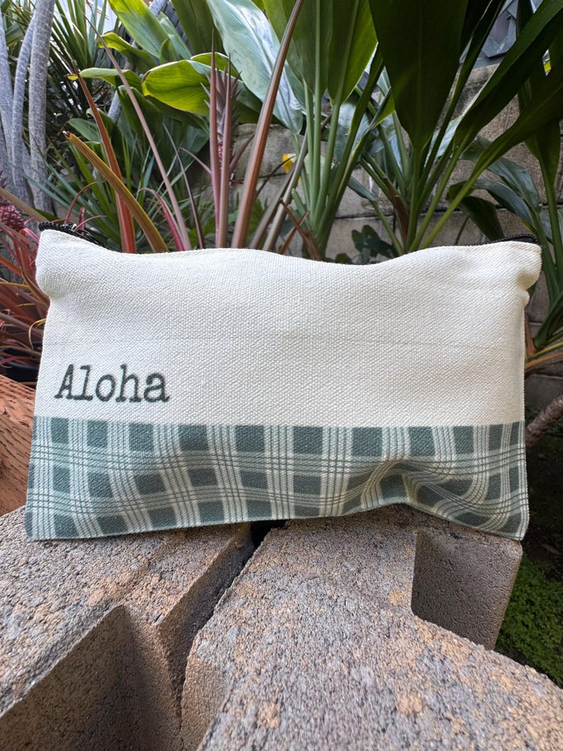 May include: Cream-colored rectangular pouch with the word "Aloha" embroidered in black. The bottom section features a green and white plaid pattern. The pouch is resting on a textured surface, with lush greenery in the background.