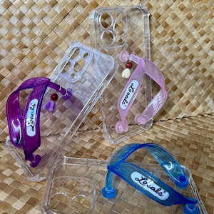 May include: Clear phone cases with colorful flip-flop style straps in purple, pink, and blue. Each strap has the word "Locals" printed on it. The cases are on a woven, light brown surface.