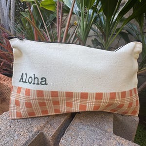 May include: Cream-colored canvas pouch with a plaid orange and brown bottom section. The word "Aloha" is printed in black on the top section. The pouch has a black zipper and is resting on a stone surface.