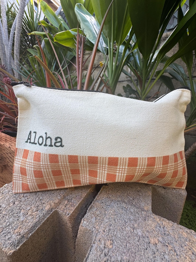 May include: Cream-colored canvas pouch with a plaid orange and brown bottom section. The word "Aloha" is printed in black on the top section. The pouch has a black zipper and is resting on a stone surface.