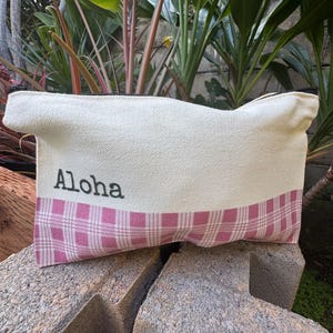 May include: A canvas pouch with the word "Aloha" printed in black. The pouch has a cream-colored top and a pink plaid bottom. The zipper is black.