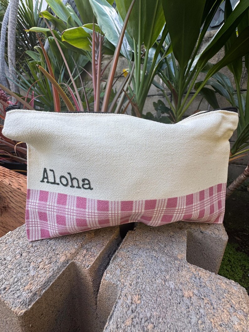 May include: A canvas pouch with the word "Aloha" printed in black. The pouch has a cream-colored top and a pink plaid bottom. The zipper is black.