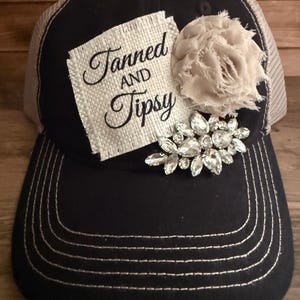 May include: A black baseball cap with a mesh back, featuring a square burlap patch with the black text Tanned AND Tipsy, a beige fabric flower with frayed edges, and a cluster of clear faceted rhinestones attached to the front panel.