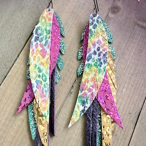 May include: A pair of colorful feather earrings. The earrings feature a mix of colors including purple, gold, green, and pink. The earrings are made of leather and have a unique, layered design. The earrings are hanging from a silver hook.
