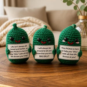 May include: Three handmade green crochet pickle figures with smiling faces and holding signs with uplifting messages. The signs read: "Life isn't always all bread and butter...", "I will always be around...", and "This PICKLE is here to help..."