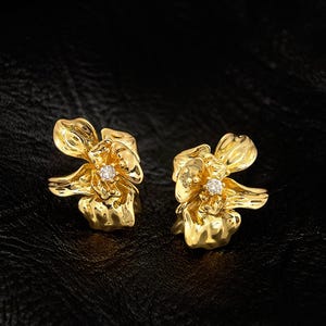 Może przedstawiać: Para złotych kolczyków w kształcie kwiatów. Każdy kolczyk ma centralny, błyszczący kamień szlachetny. Kolczyki są umieszczone na ciemnym tle, podkreślając ich skomplikowany kwiatowy wzór i metaliczny połysk.