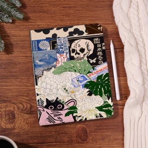 May include: A decorative notebook with a colorful Japanese-inspired design. The cover features a skull, flowers, and a cat illustration. A white stylus rests beside the notebook on a wooden surface.