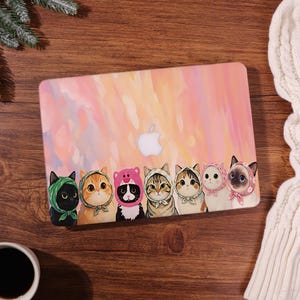 Könnte beinhalten: Eine Laptop-Hülle mit einem bunten Aquarellhintergrund und einer Reihe von Cartoon-Katzen, die verschiedene Kopfbedeckungen tragen. Die Katzen sind in verschiedenen Farben und Mustern gehalten, mit einem weißen Apple-Logo.