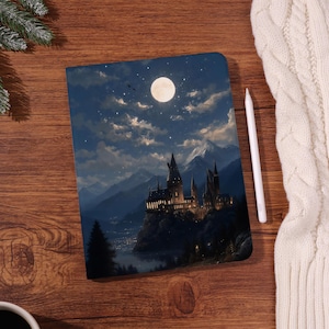 Könnte beinhalten: Ein Notizbuch mit einer nächtlichen Illustration eines Schlosses, Bergen und einem Vollmond. Das Cover ist eine detaillierte Illustration mit einem Sternenhimmel. Ein weißer Stift liegt neben dem Notizbuch auf einer Holzoberfläche.