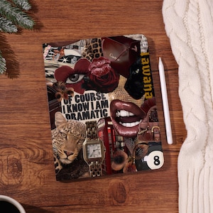 May include: A notebook with a collage design, featuring a leopard, lips, a rose, a watch, and the text "OF COURSE I KNOW I AM DRAMATIC." A white stylus is placed beside it on a wooden surface.