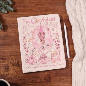 Könnte beinhalten: Ein hellrosa Notizbuch mit dem Text I'm Confident oben und I'm confident that I will get what I want unten. Der Einband zeigt einen rosa Turm, Noten, eine Krone, ein Boot und florale Illustrationen in rosa Tinte auf dem Cover.