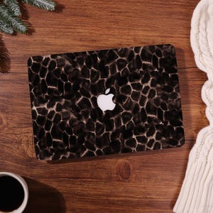 Könnte beinhalten: Eine schwarz-braun gemusterte Laptop-Hülle mit weißem Apple-Logo. Das Design der Hülle hat ein organisches, steinartiges Muster. Der Laptop liegt auf einer Holzoberfläche, mit einer Kaffeetasse und einem weißen Pullover im Hintergrund.