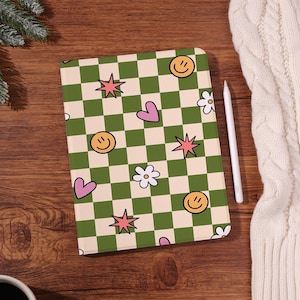 May include: A notebook with a green and cream checkered pattern, adorned with smiley faces, pink hearts, starbursts, and white flowers. A white stylus rests beside the notebook on a wooden surface.