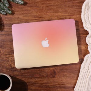 May include: A laptop computer featuring a smooth, gradient-coloured hard shell case that transitions from a soft pink at the top to a warm yellow at the bottom. The iconic white Apple logo is centred on the lid of the closed laptop device.