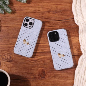 Könnte beinhalten: Zwei hellblaue Smartphone-Hüllen mit einem wiederkehrenden Muster aus kleinen schwarzen Punkten und einer einzelnen Illustration eines braun-beigen Dackels in einem cremefarbenen Pullover, mittig auf der Rückseite nahe der Kameralinse.
