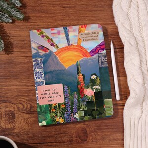 May include: A colorful journal with a collage design featuring a mountain landscape, flowers, and a setting sun. The journal has the text "Actually, life is beautiful and I have time" and "I will let myself grow even when it's hard". A white stylus rests beside it.