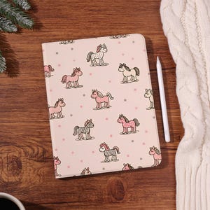 Könnte beinhalten: Eine rechteckige Tablethülle mit hellrosa Hintergrund und einem sich wiederholenden Muster aus kleinen Cartoon-Pferden in verschiedenen Rosa-, Weiß- und Brauntönen, durchsetzt mit winzigen grauen und rosa Punkten, begleitet von einem weißen Eingabestift.