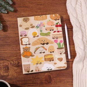 Könnte beinhalten: Ein beiges Notizbuch mit verschiedenen Illustrationen zum Thema Essen, darunter ein Croissant, ein Ei und ein Bär im Bademantel. Ein weißer Stift liegt neben dem Notizbuch. Das Notizbuch liegt auf einer Holzoberfläche.