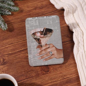 Könnte beinhalten: Ein rechteckiger Untersetzer mit einem Martini-Glas mit einer rosa Billardkugel und einem Zahnstocher. Der Untersetzer hat einen Zeitungs-Hintergrund mit dem Text "The Chicago Times". Eine Hand mit Ringen hält das Glas.