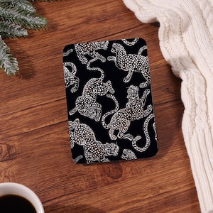 Peut inclure: Un objet rectangulaire noir avec un motif léopard blanc. Les léopards sont représentés dans diverses poses, avec des contours et des taches détaillés. L'objet est posé sur une surface en bois, avec un pull blanc et une tasse de café en arrière-plan.