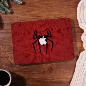 May include: Red laptop cover featuring a comic book design with a black spider and white apple logo. The cover has a glossy finish and is placed on a wooden surface. The design incorporates comic book panels.
