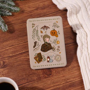 May include: A rectangular e-reader case in a light beige color featuring a central illustration of a person with a braided hairstyle surrounded by autumn-themed icons including a pumpkin, moth, lantern, book, and the text evermore autumn.