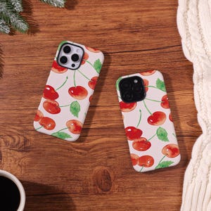 May include: Two phone cases with a cherry pattern. The cases have a white background with watercolor-style red cherries, green leaves, and thin green stems. The cases are on a wooden surface.