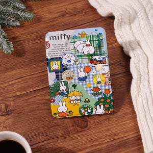 Könnte beinhalten: Rechteckige Metallbox mit der Figur Miffy, mit einer farbenfrohen Collage von Illustrationen. Das Design umfasst Miffy-Figuren, Blumen und Text. Die Box befindet sich auf einer Holzoberfläche, mit einem weißen Pullover im Hintergrund.