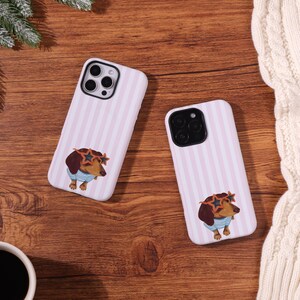 May include: Two white phone cases with pink vertical stripes and a dachshund illustration wearing star-shaped sunglasses and a blue and white striped shirt. The cases are on a wooden surface.