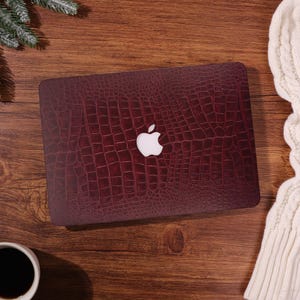May include: A burgundy laptop case with a faux alligator skin texture and a white Apple logo. The case is on a wooden surface, with a cup of coffee and a white sweater in the background. The case is designed to protect a laptop.