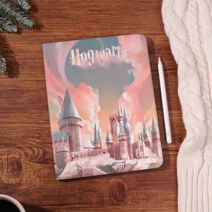 Può includere: Un quaderno con un'illustrazione colorata del castello di Hogwarts, con la parola "Hogwarts" in alto. Uno stilo bianco è appoggiato accanto al quaderno su una superficie di legno.