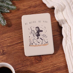 Könnte beinhalten: Ein beigefarbenes rechteckiges Etui mit dem Text "BE WEIRD. BE YOU." zeigt eine Schwarz-Weiß-Illustration eines Skeletts, das ein Einhorn reitet. Das Einhorn hat ein langes Horn und ist von kleinen Pflanzen umgeben.