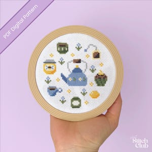 May include: A cross-stitch design featuring a blue teapot, teacup, honey jar, and other tea-related elements. The design is set in a round wooden embroidery hoop. The text "PDF Digital Pattern" is visible in the upper left corner.