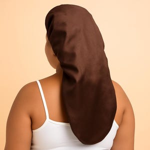 May include: A dark brown satin bonnet, designed to protect hair. The bonnet is long and covers the head and neck. The person in the image is wearing a white tank top. The background is a solid, light peach color.