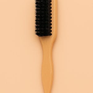May include: A wooden brush with a handle and a rectangular head. The head features a row of black bristles. The brush is made of light-colored wood and is designed for grooming.