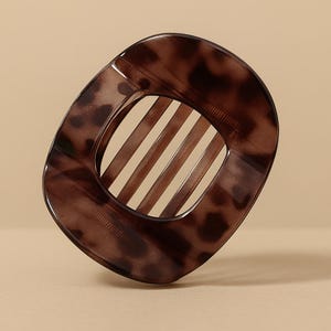 Op de afbeelding: Een bruine haarclip met een schildpadpatroon. De clip heeft een rechthoekige vorm met afgeronde hoeken en een reeks parallelle staven in het midden. De clip is gemaakt van een doorschijnend materiaal.