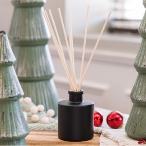May include: A black essential oil diffuser with wooden reeds, set against green Christmas tree decorations and red baubles. The cylindrical diffuser sits on a wooden surface, creating a festive holiday scene.