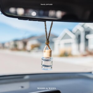 May include: A clear glass travel diffuser hanging from a car's rearview mirror. The diffuser has a wooden top and a beige rope. The text "TRAVEL DIFFUSER" is above the mirror, and "WOODS & WAVES" is below.