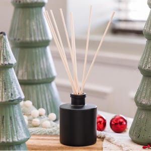 May include: A black essential oil diffuser with wooden reeds, placed in front of green Christmas tree ornaments and red baubles. The diffuser is cylindrical and rests on a wooden surface, creating a festive holiday scene.