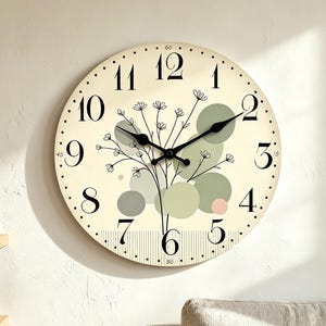 May include: A round wall clock with a cream-colored face featuring black numerals from 1 to 12. The design includes a central illustration of black line-art flowers, overlapping sage green and pale pink circles, and a vertical striped pattern at the bottom.