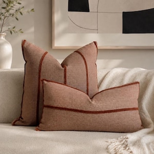 May include: Two terracotta-colored throw pillows with a textured fabric finish. The square pillow features vertical piping, while the rectangular lumbar pillow displays a single horizontal line of raised, dark orange piping across the center.