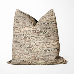 May include: Decorative throw pillow with a beige, brown, and black abstract pattern. The pillow features a textured design with a mix of organic shapes and lines, creating a visual depth. The pillow is a square shape.