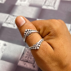 May include: Two silver-colored chevron rings with a beaded design are worn on a finger. The rings have a V-shape, with the beaded pattern forming the top of the V. The rings are on a hand with brown skin.