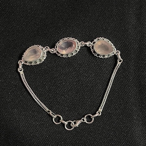 May include: A silver bracelet featuring three faceted, pale pink rose quartz gemstones set in ornate, scalloped silver bezels. The bracelet is constructed with a delicate herringbone chain and includes a lobster claw clasp with adjustable jump rings.