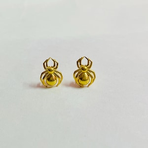 May include: Two gold-colored spider-shaped stud earrings. Each earring features a rounded body and detailed legs and head. The earrings are displayed on a white surface, showcasing their intricate design and metallic sheen.