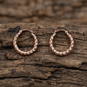 Op de afbeelding: Een paar roségouden oorringen met een gedraaide touwtextuur. Elke cirkelvormige oorbel is aan de bovenkant voorzien van een scharnierend sluitingsmechanisme, wat zorgt voor een doorlopend, afgerond silhouet met een gepolijste, metalen afwerking.