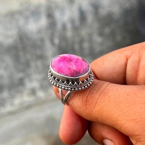 May include: A ring featuring a large, oval-shaped, vibrant pink gemstone set in a silver-toned metal band. The setting displays a decorative, beaded texture along the edge of the bezel, while the band itself consists of a split, polished metal shank.
