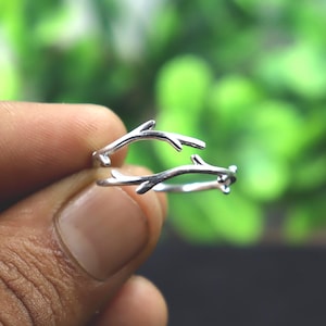 May include: A silver ring with a twig design. The ring has two branches that curve and meet, forming an open design. The ring is displayed against a blurred green backdrop.